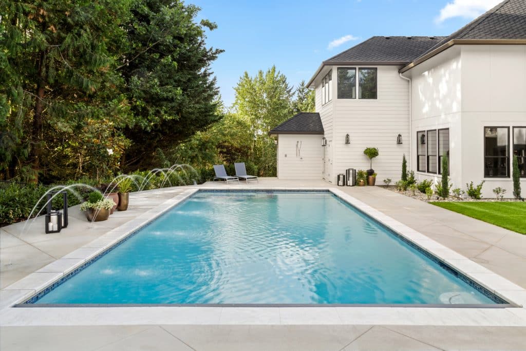 Spacious backyard pool with water features beside a modern white home and landscaped yard.
