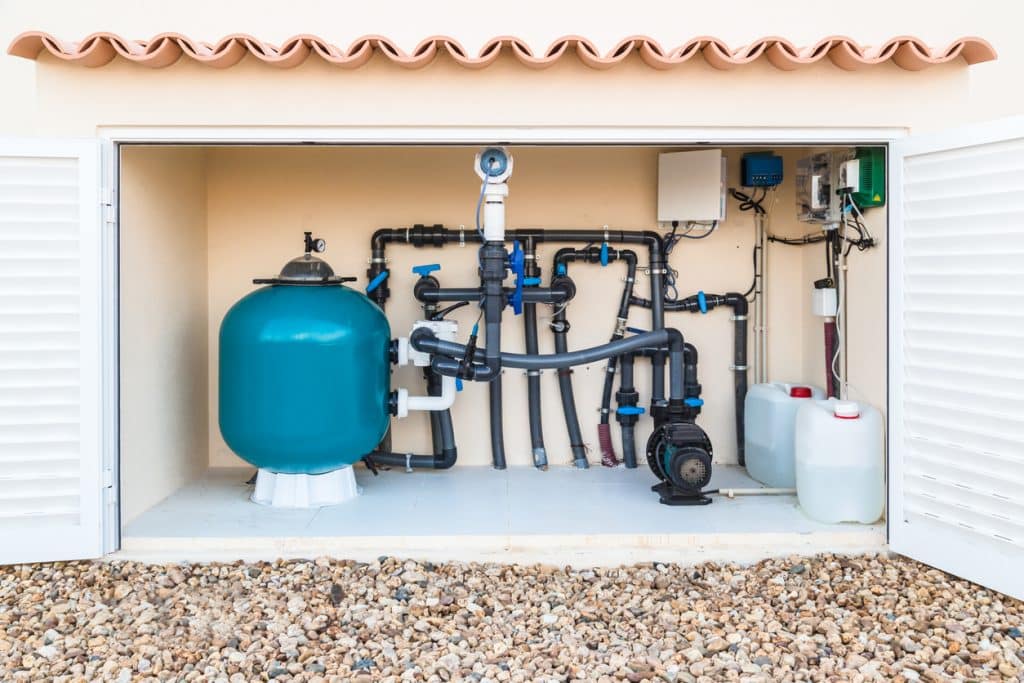 A pool equipment enclosure containing a large filter tank, pump, pipes, valves, hoses, and chemical containers for swimming pool filtration and maintenance.
