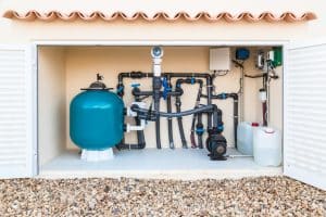 A pool equipment enclosure containing a large filter tank, pump, pipes, valves, hoses, and chemical containers for swimming pool filtration and maintenance.