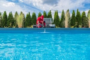 A pool technician in red workwear kneels beside a swimming pool, inspecting or servicing the pool equipment on a sunny day.