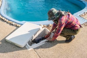 A pool technician repairs a swimming pool pump inside an open equipment compartment beside a clear blue swimming pool.
